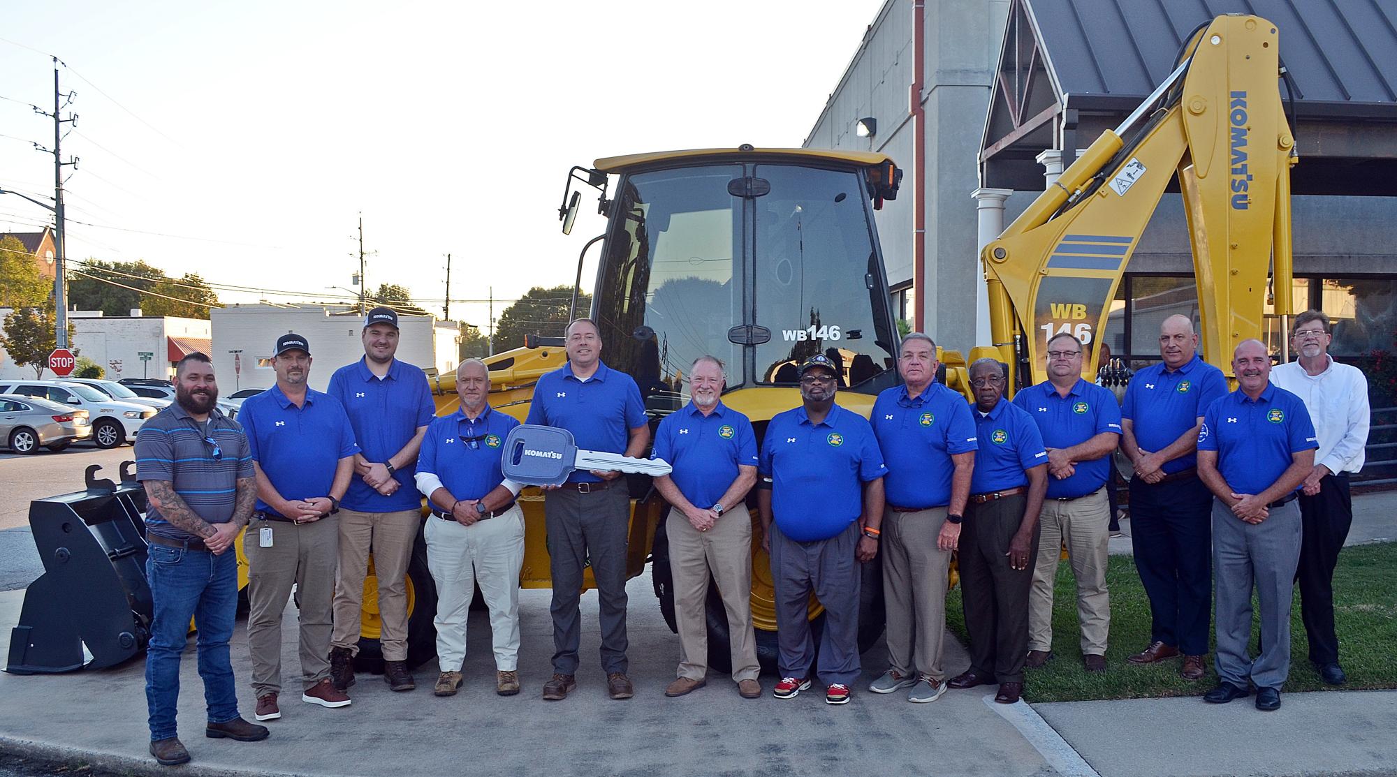 Alt text: A group poses beside the refurbished yellow Komatsu loader/backhoe outside a building. Most wear matching blue polo shirts with county emblems. An oversized ceremonial Komatsu key is held near the center of the group.